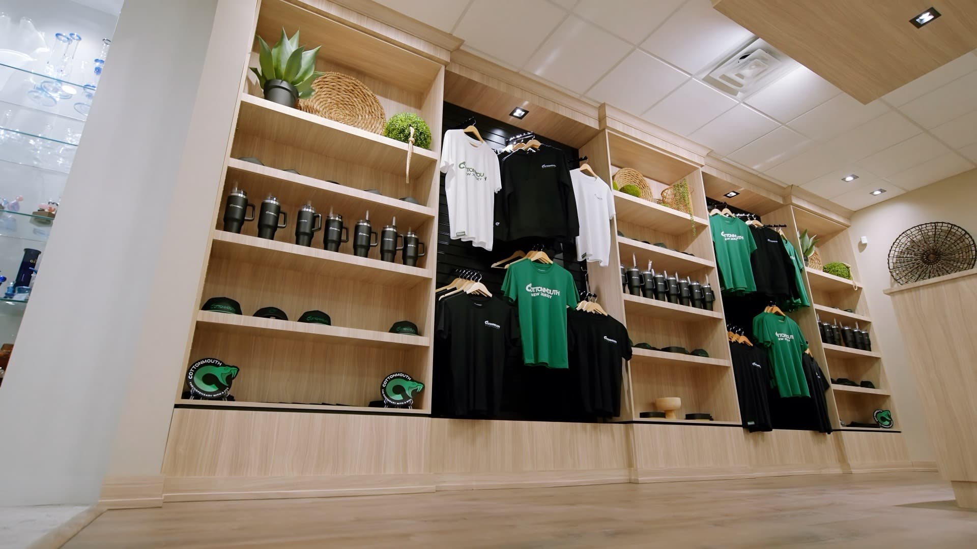 The Cottonmouth merch wall inside the Runnemede dispensary, with branded tees, hats, and tumblers on oak shelving